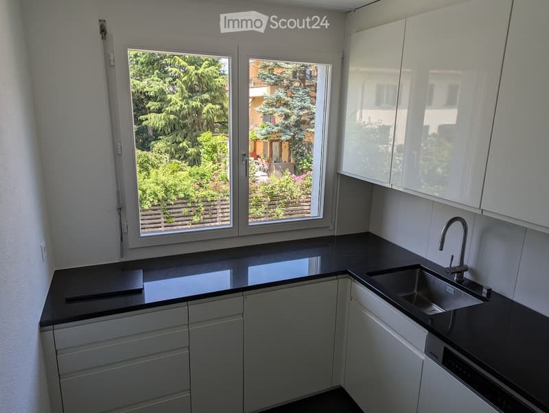 Modern kitchen with white cabinets, black countertops, and a stainless steel sink. Large windows offer a view of a garden with trees and flowers.