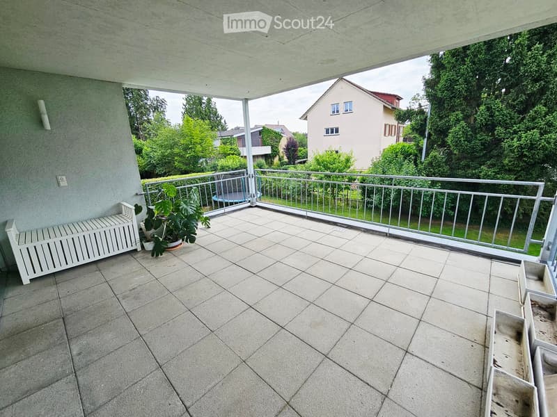 Balcony with tiled floor, metal railing, bench, and potted plants, overlooking a residential area with houses and greenery.