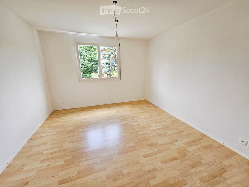 Empty room with light wooden flooring and white walls, featuring a single window with a view of greenery outside.