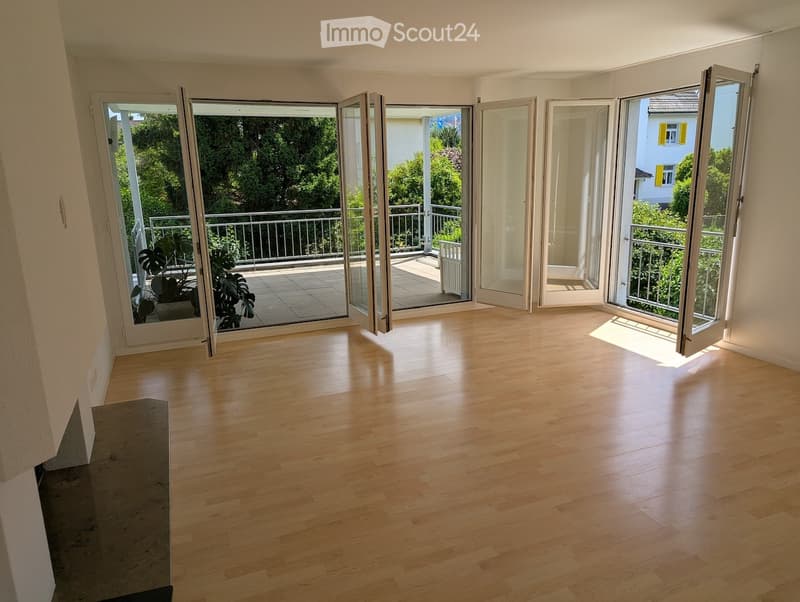 Empty room with light wooden flooring, large glass sliding doors leading to a balcony with a metal railing and outdoor plants.