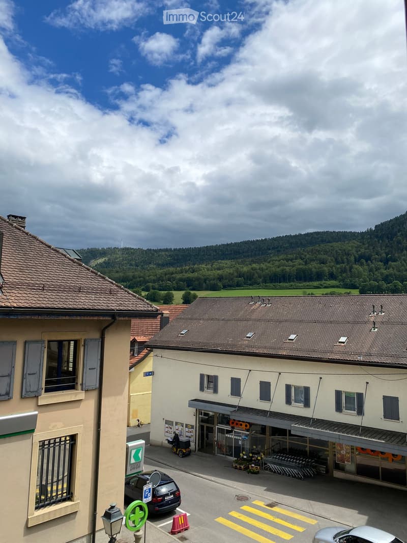 A two-story building with a tiled roof and multiple windows, situated in a rural area with a view of green hills and trees in the background. There is a parking area in front with a few cars and a small shop entrance visible.