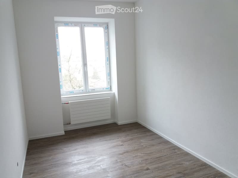 Empty room with light wooden flooring, white walls, and a single window with a radiator below it.