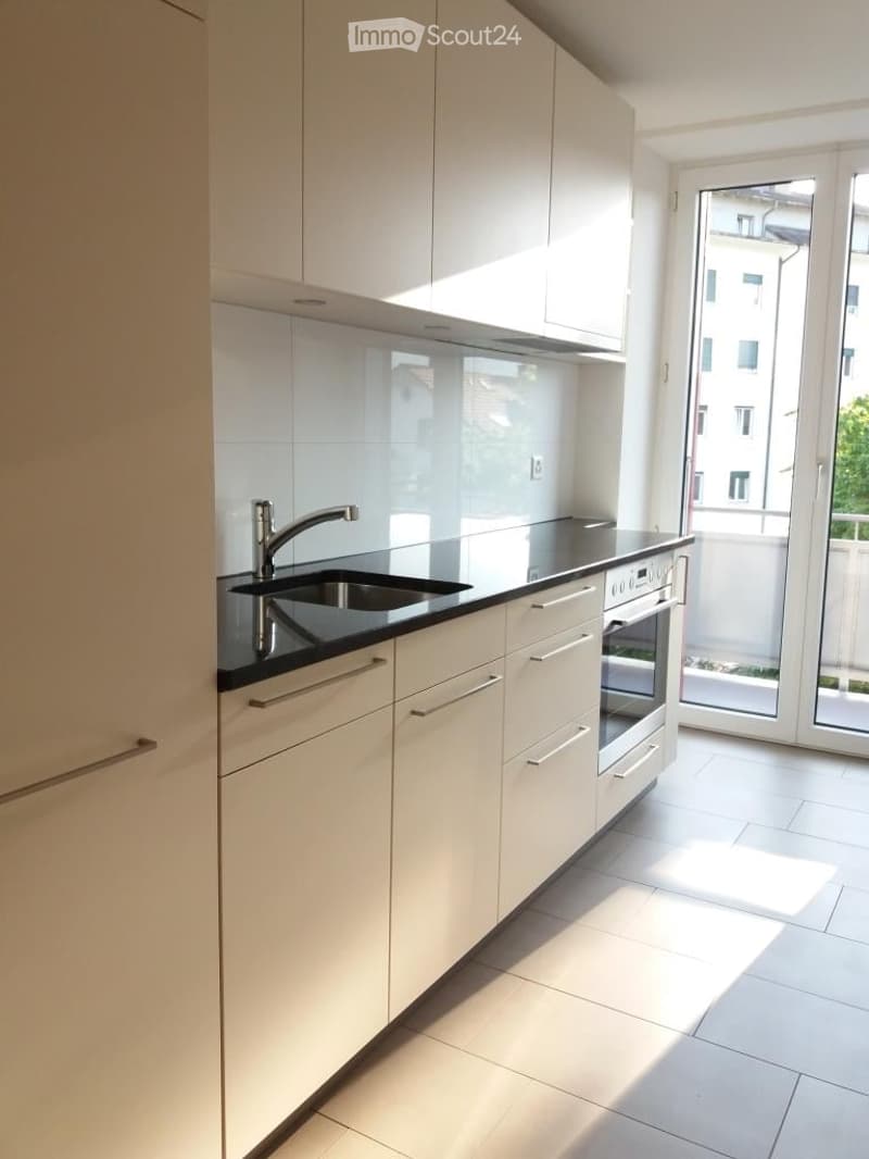 Modern kitchen with white cabinets, black countertops, a sink, and an oven. Large windows offer a view of the outside.