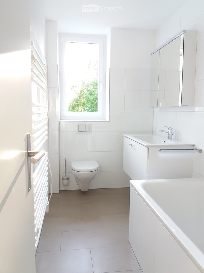 Modern bathroom with white tiles, a toilet, a bathtub, a sink with a faucet, a mirror, and a cabinet.