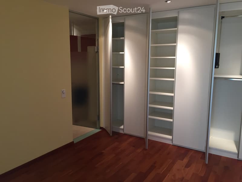 Empty room with wooden flooring, white built-in closet with open shelves, and a mirrored sliding door leading to another space.