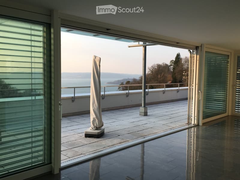 Modern balcony with large glass doors, tiled floor, covered umbrella stand, and scenic view of water and hills.
