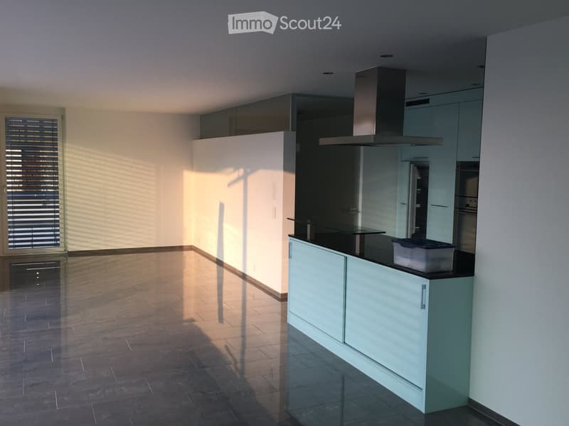 Modern kitchen with light grey cabinets, a central island, and stainless steel appliances including a range hood, refrigerator, and oven. The space features a tiled floor and large windows with blinds allowing natural light to fill the room.