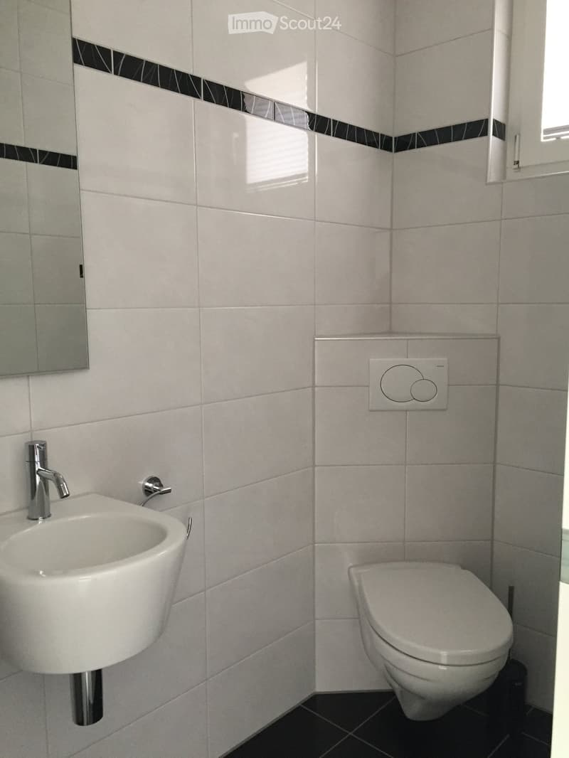 Modern bathroom with white tiles, a wall-mounted sink with a chrome faucet, and a toilet. The walls feature a decorative black and white border near the top.