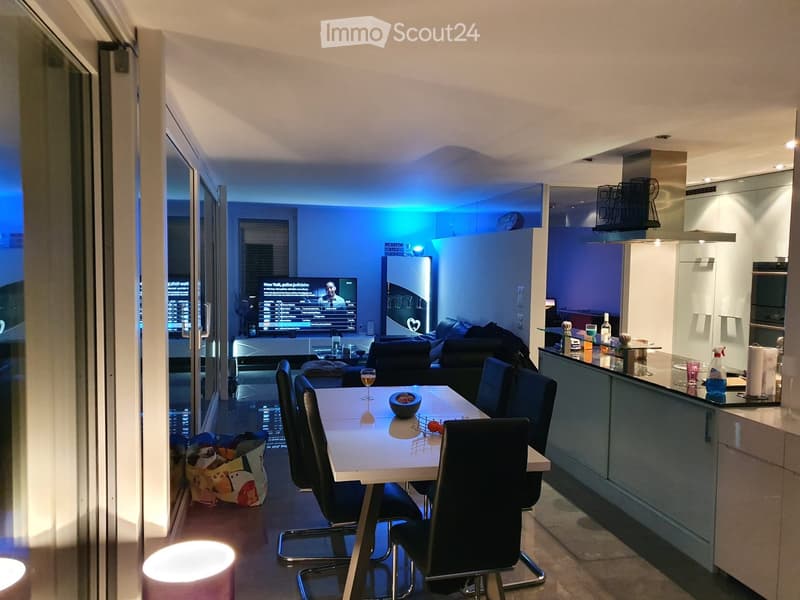 Modern open-plan living and dining area with a sofa, dining table, chairs, and a kitchen counter. The space features large windows with a city view and ambient blue lighting.