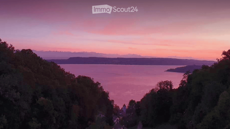 Panoramic view of a lake at sunset, surrounded by trees and distant hills.