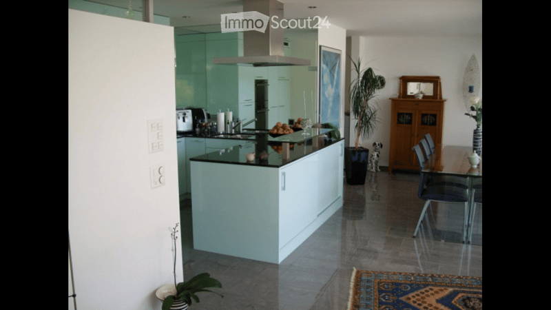 Modern kitchen with a central island, sleek white cabinets, and a glass countertop. The space includes a sink, faucet, fridge, oven, and a small dining area with a table and chairs. Decorative elements include a plant, a mirror, and an art piece on the wa