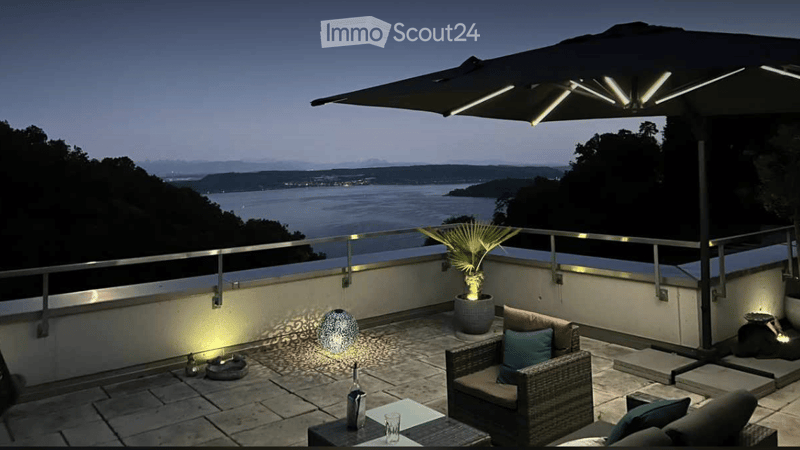 Balcony with a view of the sea, featuring a large umbrella, wicker chairs, a small table, and a potted plant.