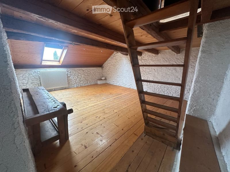 Empty attic space with wooden floors, exposed wooden beams, stone walls, a small window, and a radiator. No furniture or equipment present.