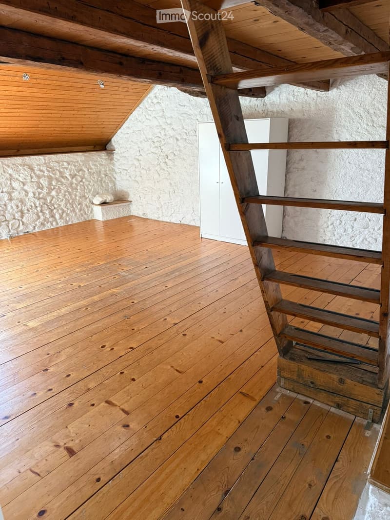 An attic space with wooden floors and exposed wooden beams, featuring a staircase leading upwards and a built-in shelf on the wall.