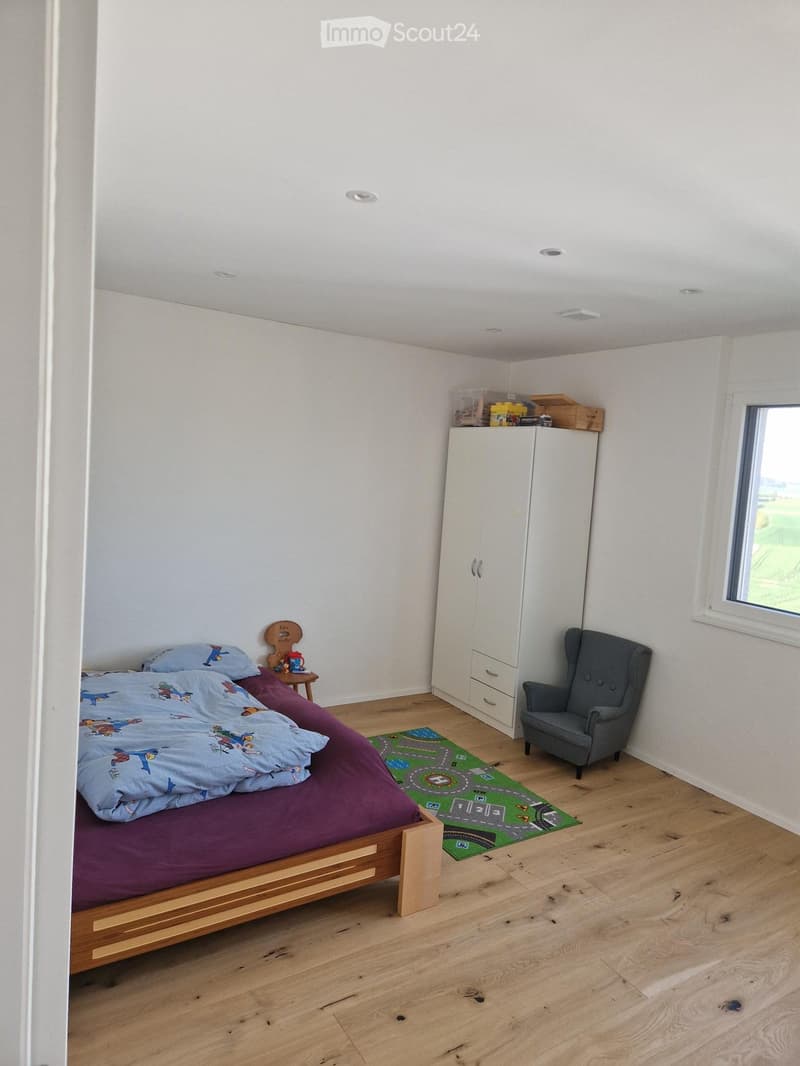 Bright bedroom with wooden flooring, a bed with colorful bedding, a wardrobe, a small table with a lamp, a grey armchair, and a green play mat on the floor.