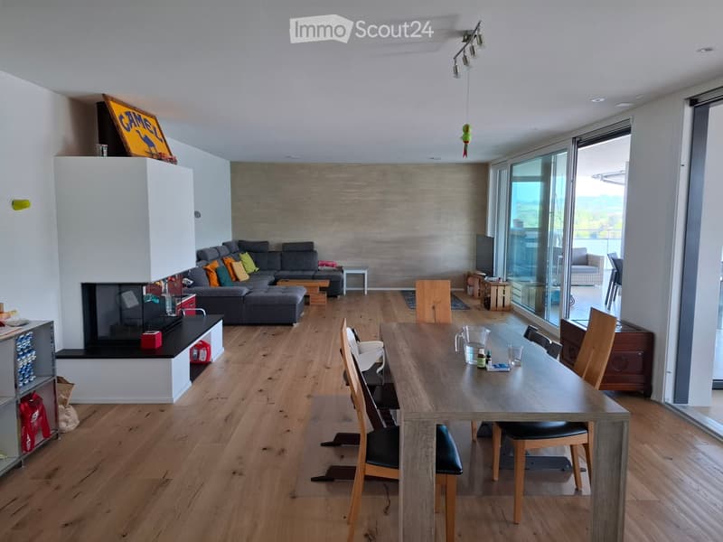 Open-plan living and dining area with wooden flooring, a grey sectional sofa, a fireplace, a dining table with chairs, and large windows offering a scenic view.