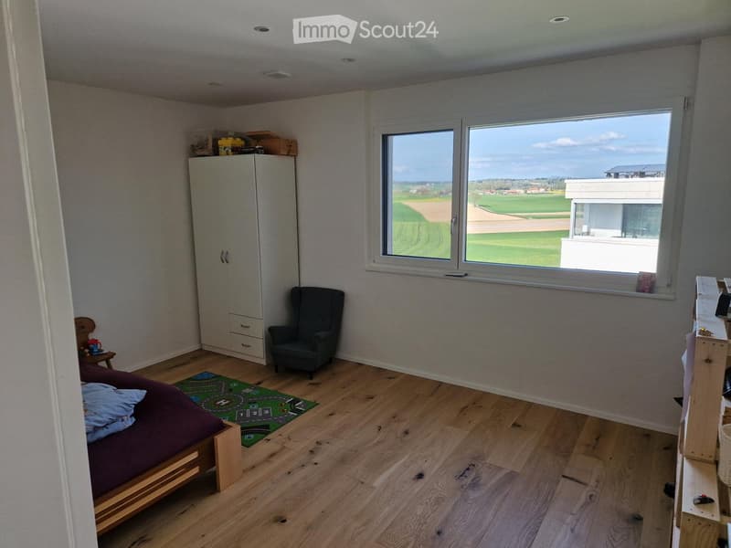 Bright bedroom with wooden flooring, large window overlooking a green field, bed with purple bedding, wardrobe, armchair, small table, and toy on the floor.