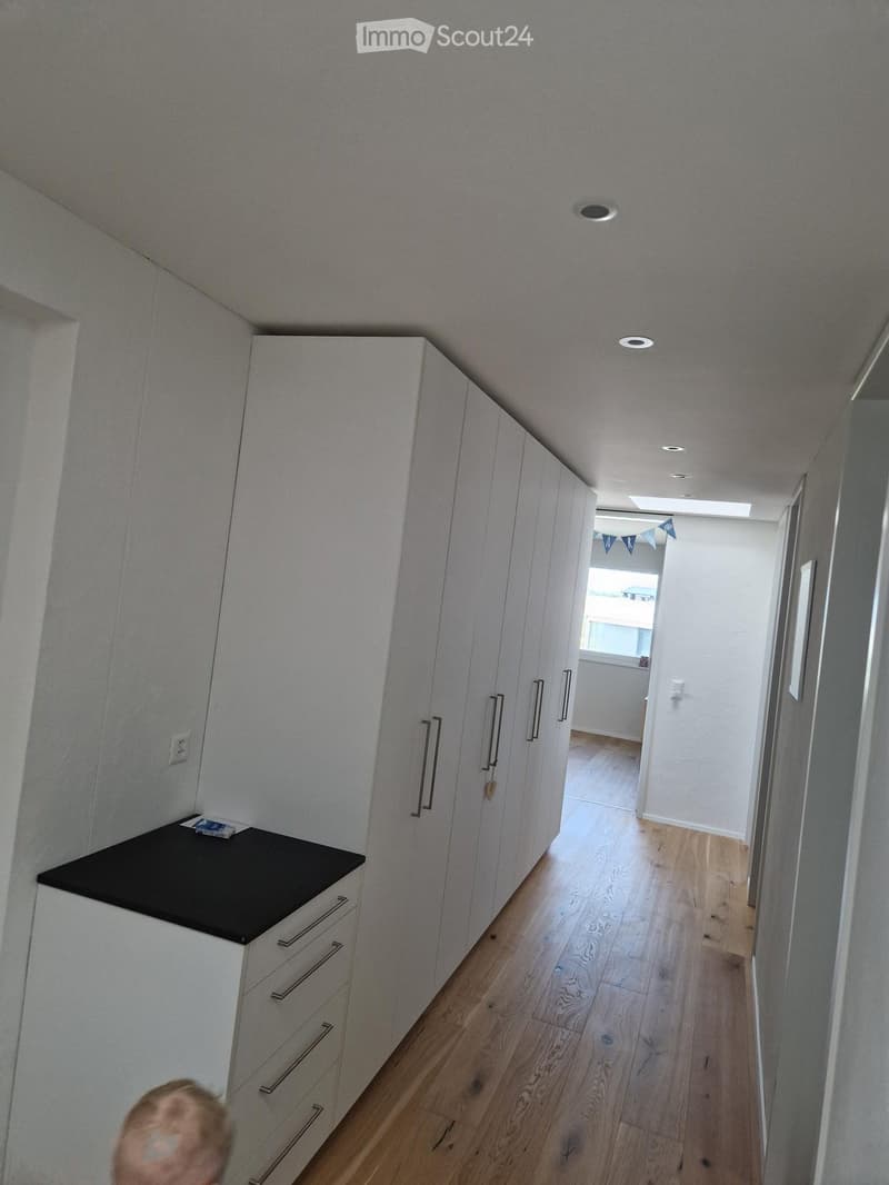 A modern hallway with light wooden flooring, white walls, built-in white cabinets with silver handles, and recessed ceiling lighting. A small black countertop with a book is visible on the left side.