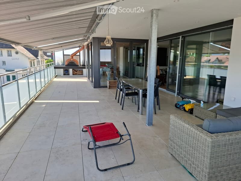 Balcony with tiled floor, glass railing, outdoor furniture including a table, chairs, a wicker sofa, and a red lounge chair.