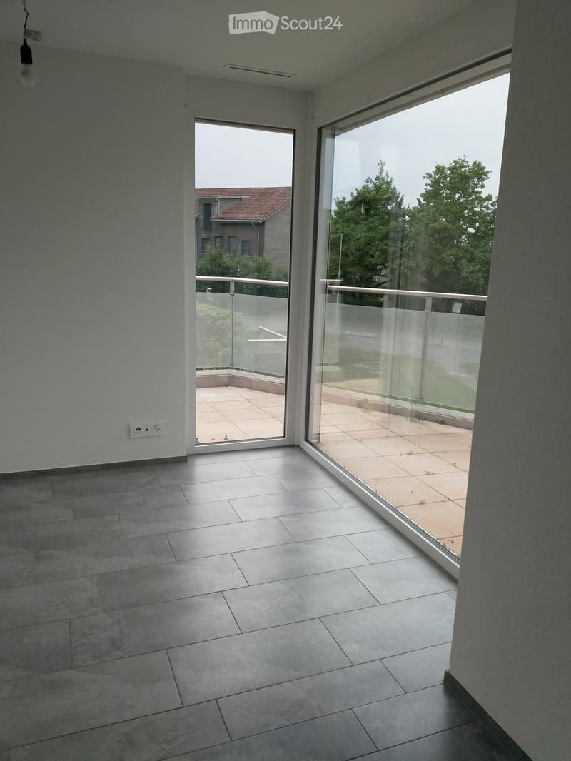 Modern interior space with large glass doors leading to a tiled balcony area, featuring a view of greenery and neighboring buildings.