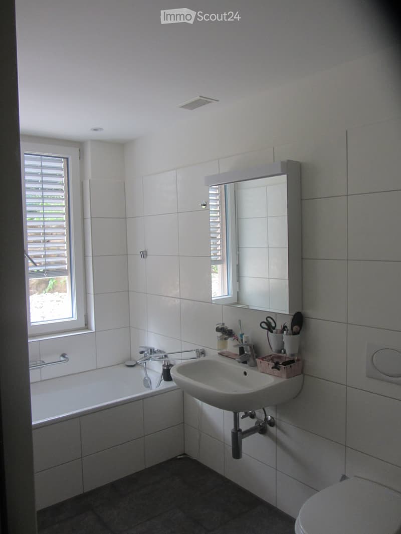 Bathroom. Modern bathroom with white tiled walls, a bathtub, a sink, a mirror, and a toilet. The room features a window with blinds and a small shelf holding toiletries.