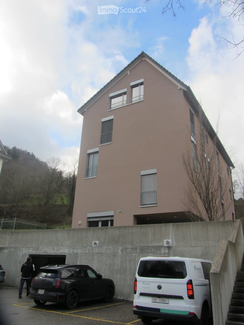 Building. Two-story residential building with light pink facade, multiple windows, and a garage entrance. Two cars are parked in front of the building.
