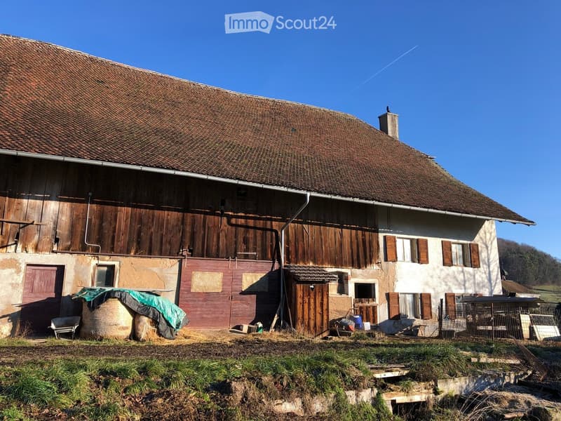 A traditional old house made of stone walls and wooden structure, with several doors, windows, and a chimney on the right side. A roof over the house is brown and old, and the house seems to be situated on a vast grassy field. Two hay bales are on the fro