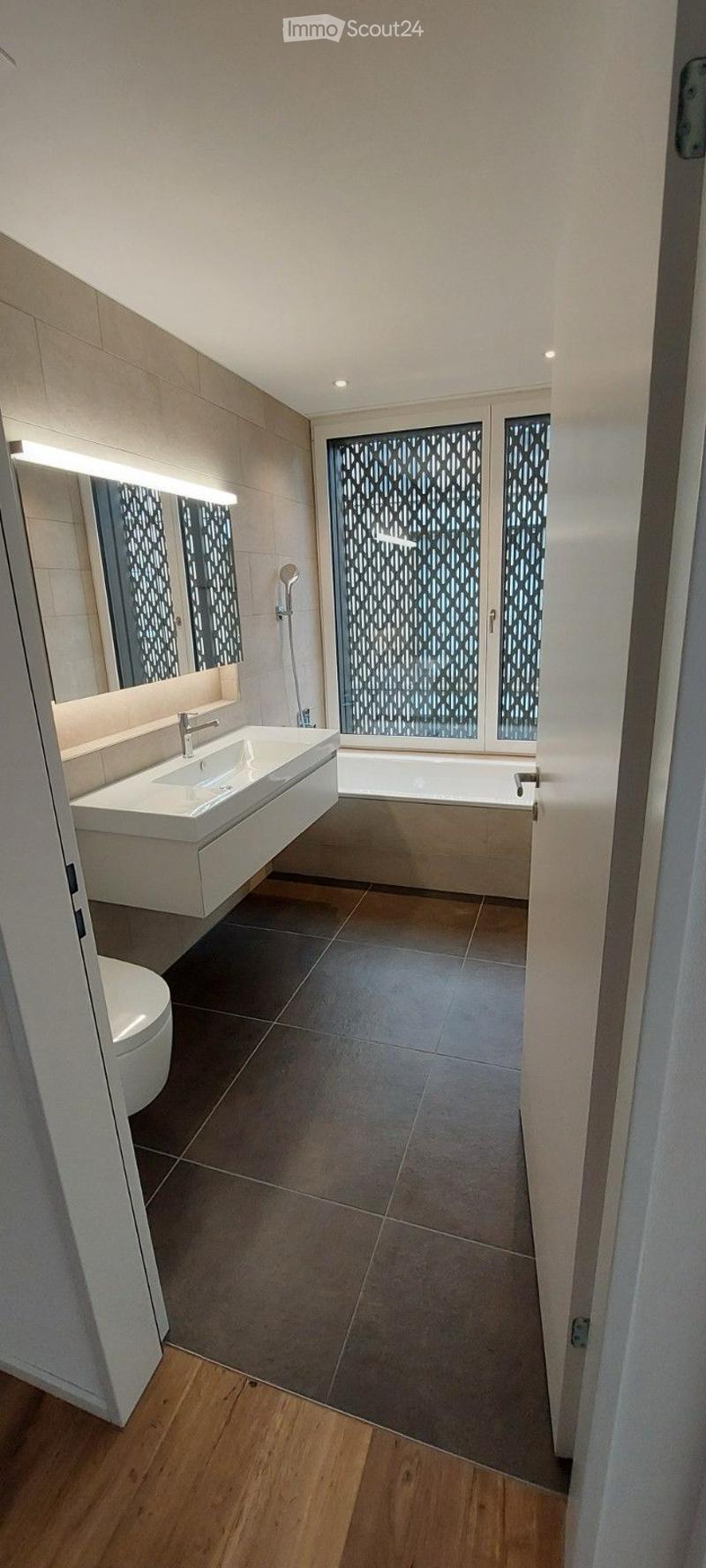 Modern bathroom with a white sink, bathtub, toilet, and large mirror. The space features dark tiled flooring and light-colored walls.