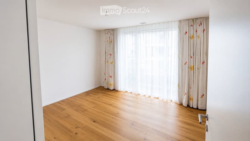 Empty room with wooden flooring, white walls, and large window with floral curtains.