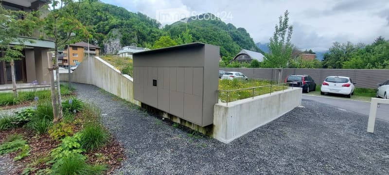 Outdoor area with a parking lot, modern building, greenery, and a raised ground floor entrance.