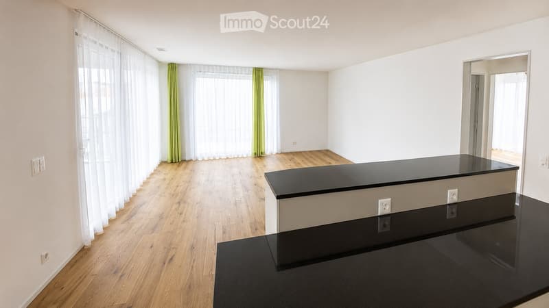 Empty room with light wooden flooring, white walls, large windows with white and green curtains, and a black countertop in the corner.