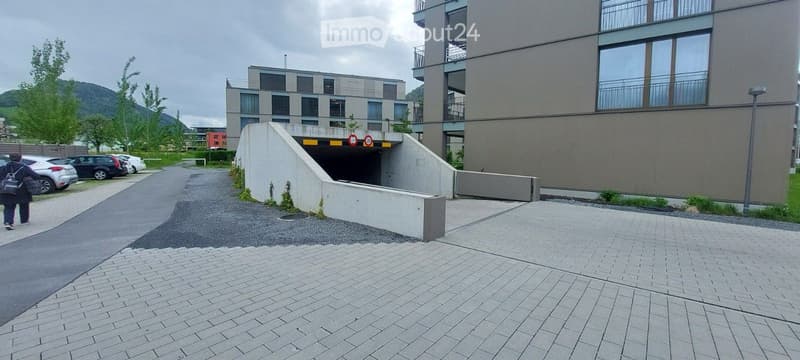 Modern multi-story apartment building with a covered garage entrance and adjacent parking area.
