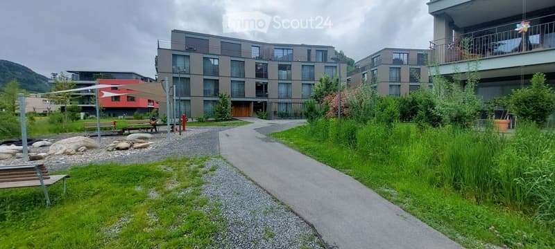 Modern residential complex with a landscaped garden area, featuring a paved pathway, benches, and greenery.