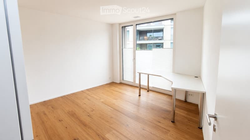 A small, unfurnished room with wooden flooring, white walls, and a large window with blinds. A small table and a stool are present.