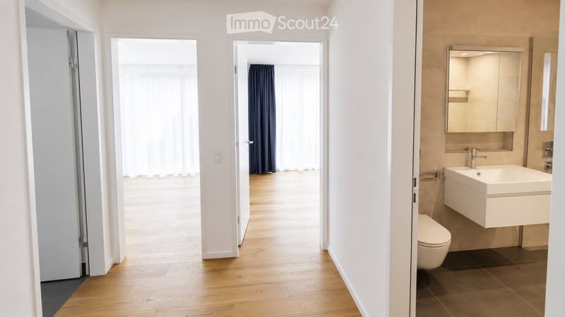 A modern, well-lit hallway with wooden flooring and white walls, leading to a bathroom with a sink, mirror, and toilet.