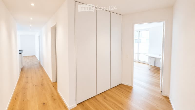 A clean, modern hallway with light wooden flooring, white walls, and a sliding door leading to a room with a small table and chairs.