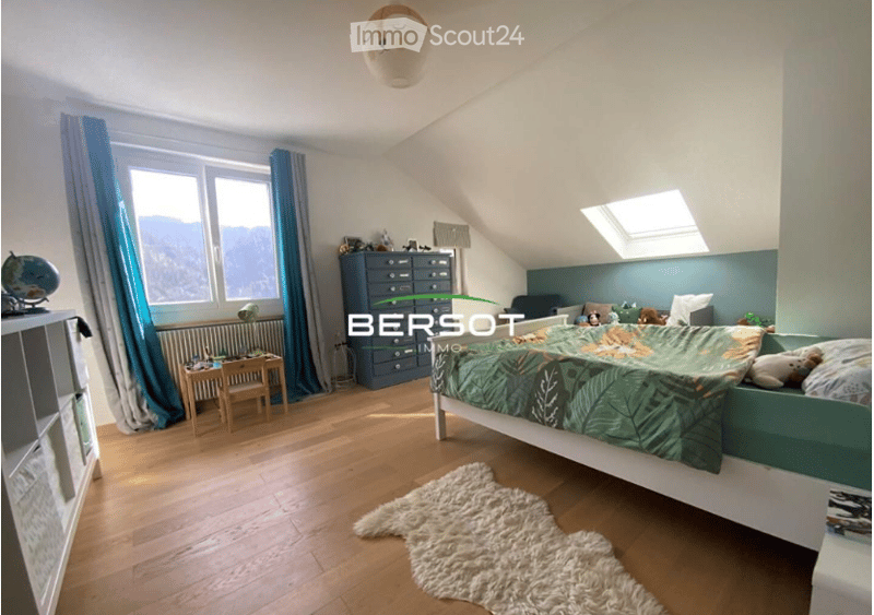 Bright bedroom with wooden flooring, large window with blue curtains, bed with green and yellow bedding, nightstand, small table, cabinet, and decorative items including a globe and toys.