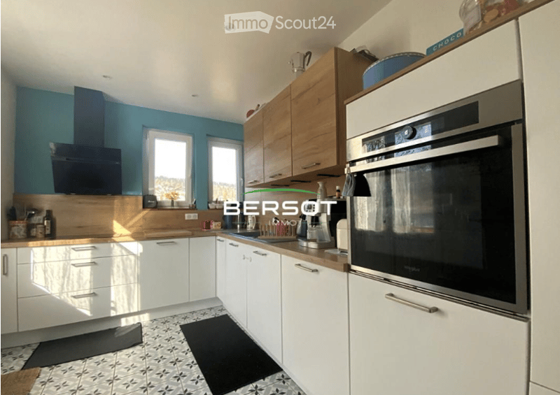 Modern kitchen with white and light wood cabinets, stainless steel oven, island, and countertop. Features a blue accent wall and tiled floor.