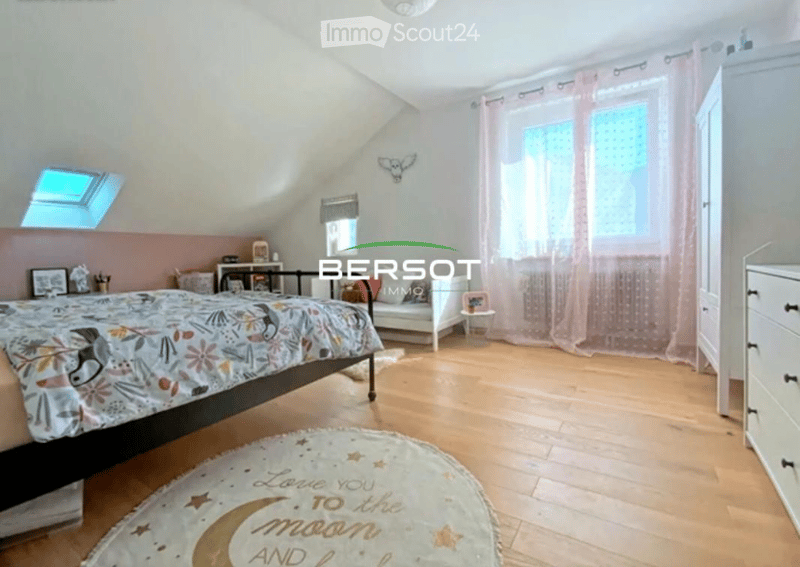 Cozy attic bedroom with wooden flooring, slanted ceilings, large windows with pink curtains, a bed with a colorful duvet, a nightstand, a mirror, a small chair, and a white wardrobe.
