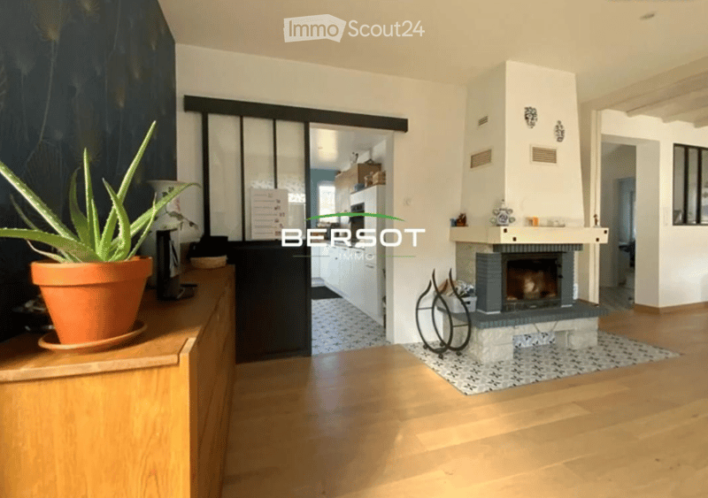 Bright living room with wooden flooring, fireplace, potted plant, and open shelving area visible in the background.