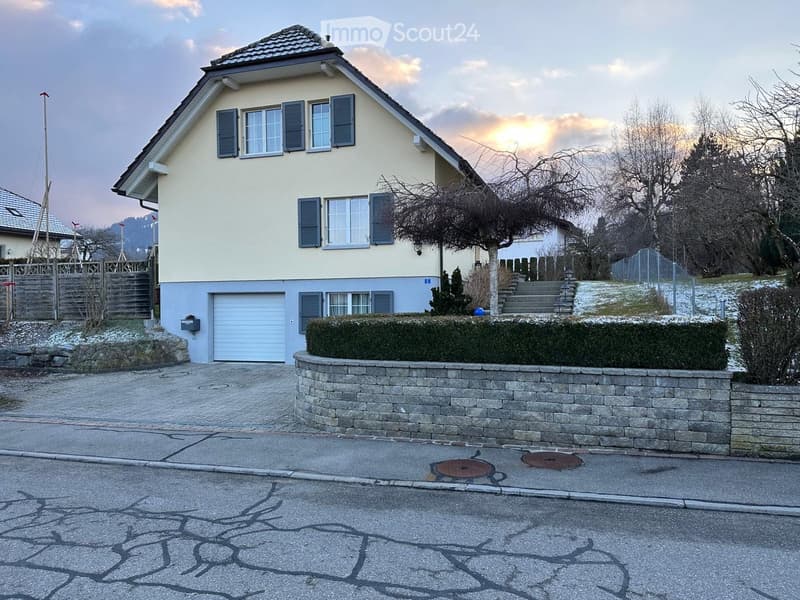Two-story house with beige exterior, blue shutters, garage, stone wall, and steps leading to entrance.