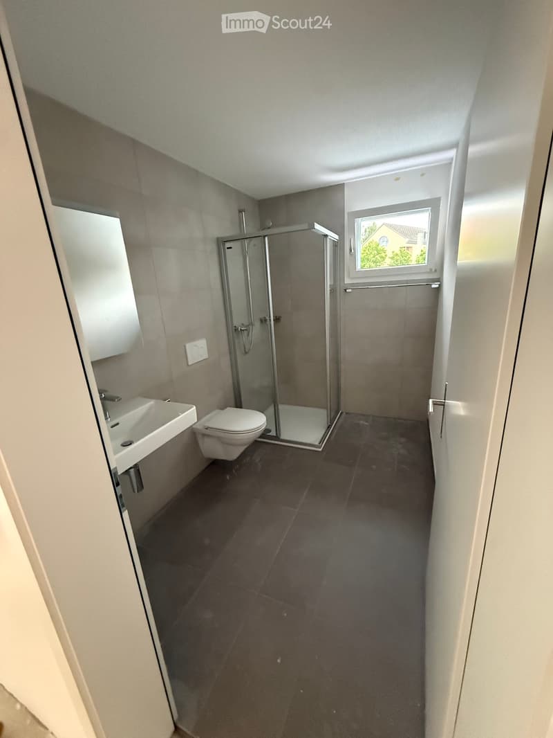 Modern bathroom with dark tiled floor, white sink, toilet, and glass-enclosed shower. A window provides natural light.