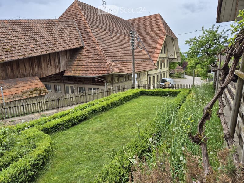 Traditional wooden house with red-tiled roof, surrounded by a well-maintained garden with hedges and flowers, and a wooden fence.