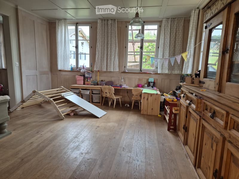 Brightly lit room with wooden flooring, large windows, and various children's furniture including a desk, chairs, a toy slide, and storage bins.