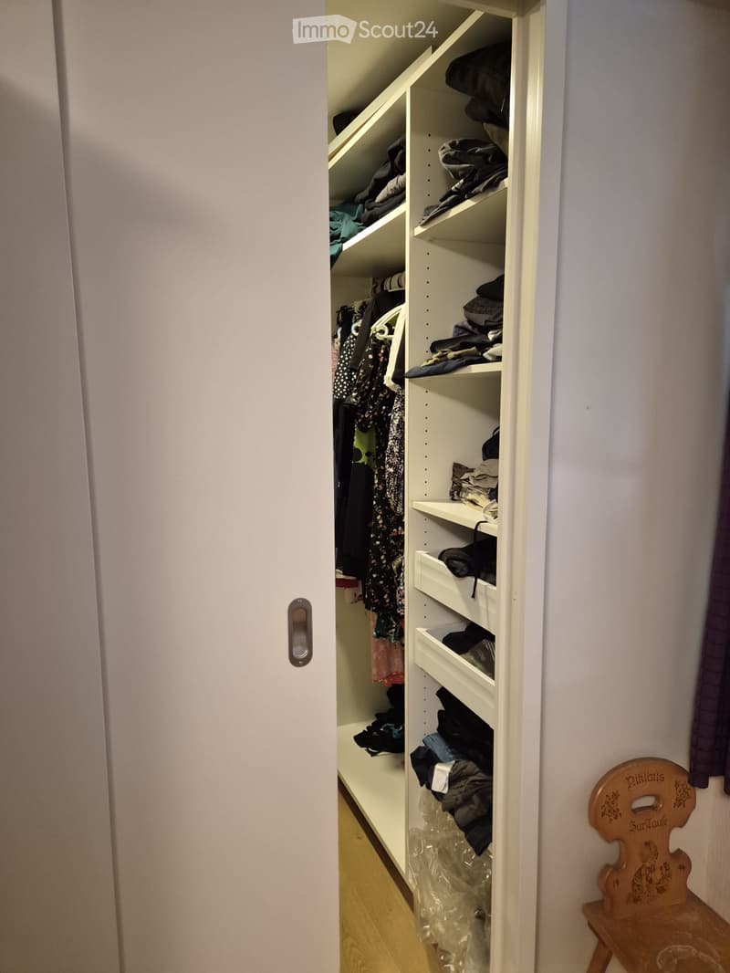 A well-organized closet with white shelves filled with folded clothes and items, a small wooden chair with a decorative backrest is placed next to the closet.