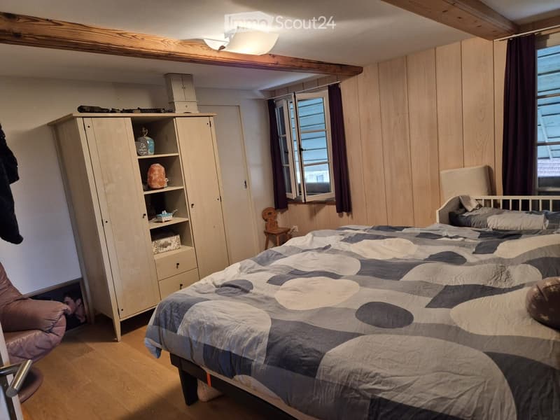 Cozy bedroom with wooden walls and ceiling, featuring a double bed with a grey and white patterned duvet, a tall wardrobe with shelves, a small wooden stool, and a pink armchair.