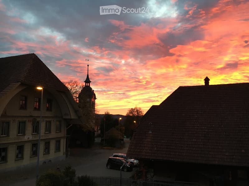 Village scene at sunset with traditional houses, a church tower, and parked cars.