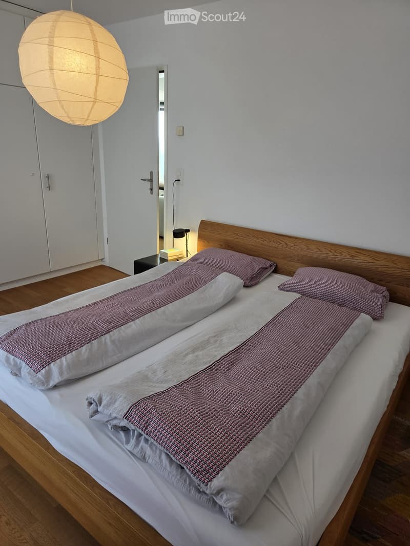 Bedroom. A neatly made double bed with a wooden headboard, pillows, and a patterned duvet, positioned on a wooden floor. A pendant light hangs from the ceiling, and a closet is visible in the background.