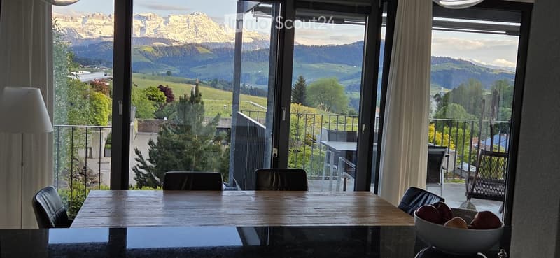 Furnished Room. Dining area with a wooden table, black chairs, and a bowl of fruit, overlooking a scenic mountain and valley view through large windows and a balcony.