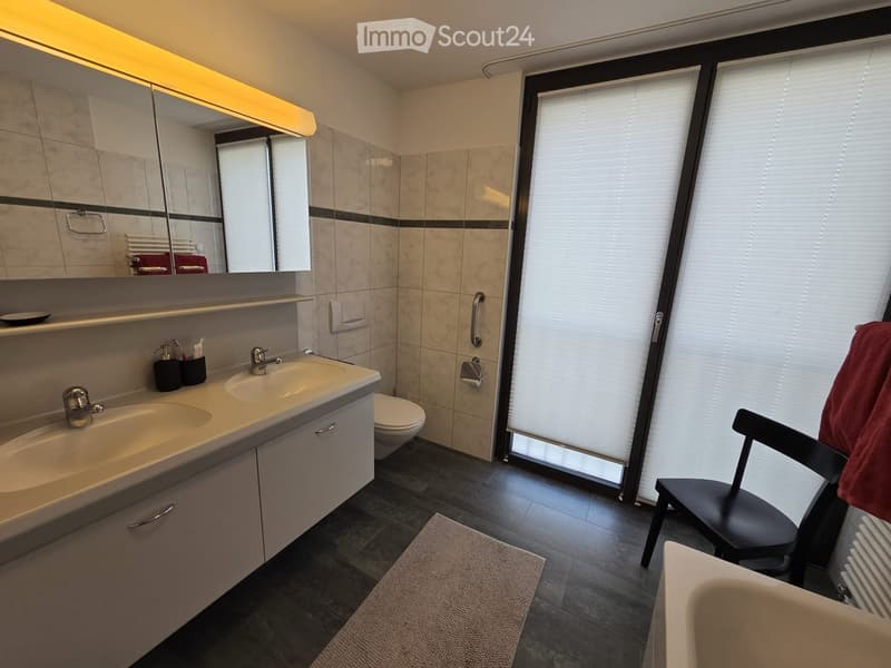 Bathroom. Modern bathroom with double sink, toilet, large mirror, and a window with blinds. Includes a chair with a towel draped over it and a small rug on the floor.